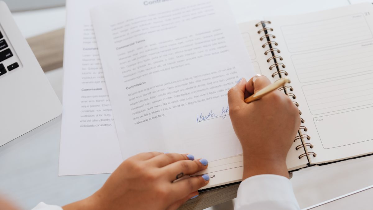 Two people signing a contract document with a pen beside a notebook, representing the one-page agreement a builder sends before starting.